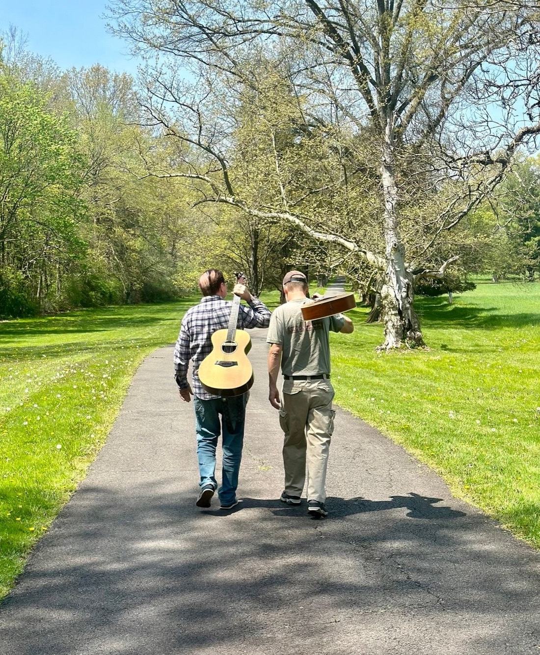 Chris Bukata and Paul Heitmann walking down a tree-lined path carrying acoustic guitars